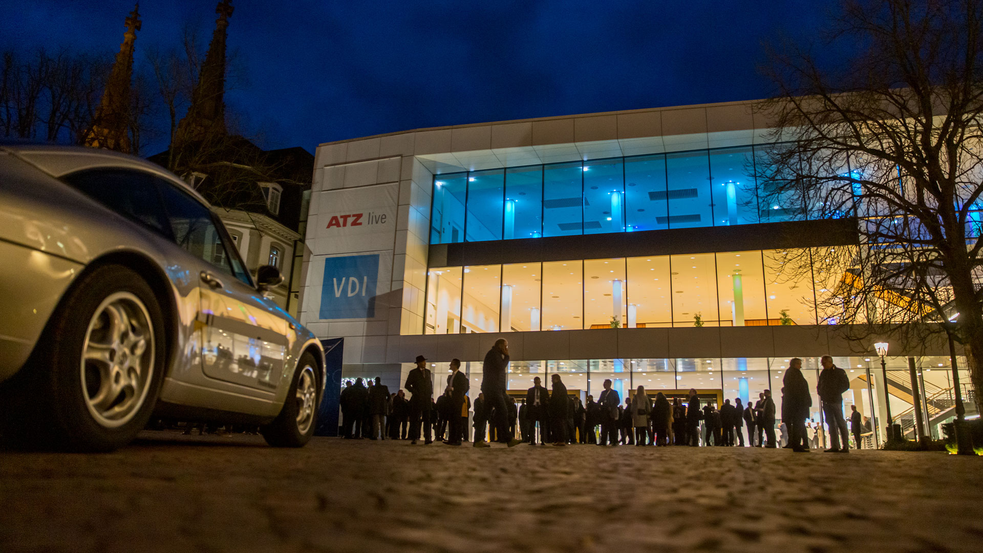 Viele Konferenzteilnehmer vor dem Kongresshaus am Abend. Im Vordergrund ein Ausstellungsfahrzeug. 